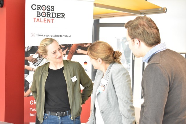 Eine Frau unterhält sich mit einer weiteren Frau und einem Mann vor einem Banner des Projekts "Cross Border Talent"