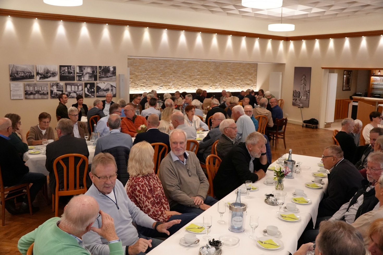 In einem großen Saal sitzen an mehreren langen Tischen viele ältere Personen und unterhalten sich miteinander.