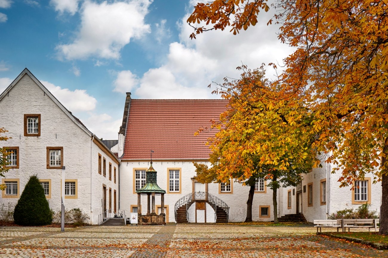 Der ehemalige Adelshof Falkenhof in Rheine (Bildquelle: Münsterland e.V., Philipp Fölting) Historischer Innenhof mit gepflastertem Boden, umgeben von weißen Gebäuden mit roten Ziegeldächern und braun umrahmten Fenstern. In der Mitte steht ein überdachter Brunnen mit grünem Kupferdach. Rechts und links des Platzes stehen Kastanienbäume mit herbstlich gefärbtem Laub in Gelb-, Orange- und Rottönen. Eine Treppe mit geschwungenem Geländer führt zur Eingangstür eines zentralen Gebäudes. Der Himmel ist teils bewölkt mit blauem Himmel und weißen Wolken.