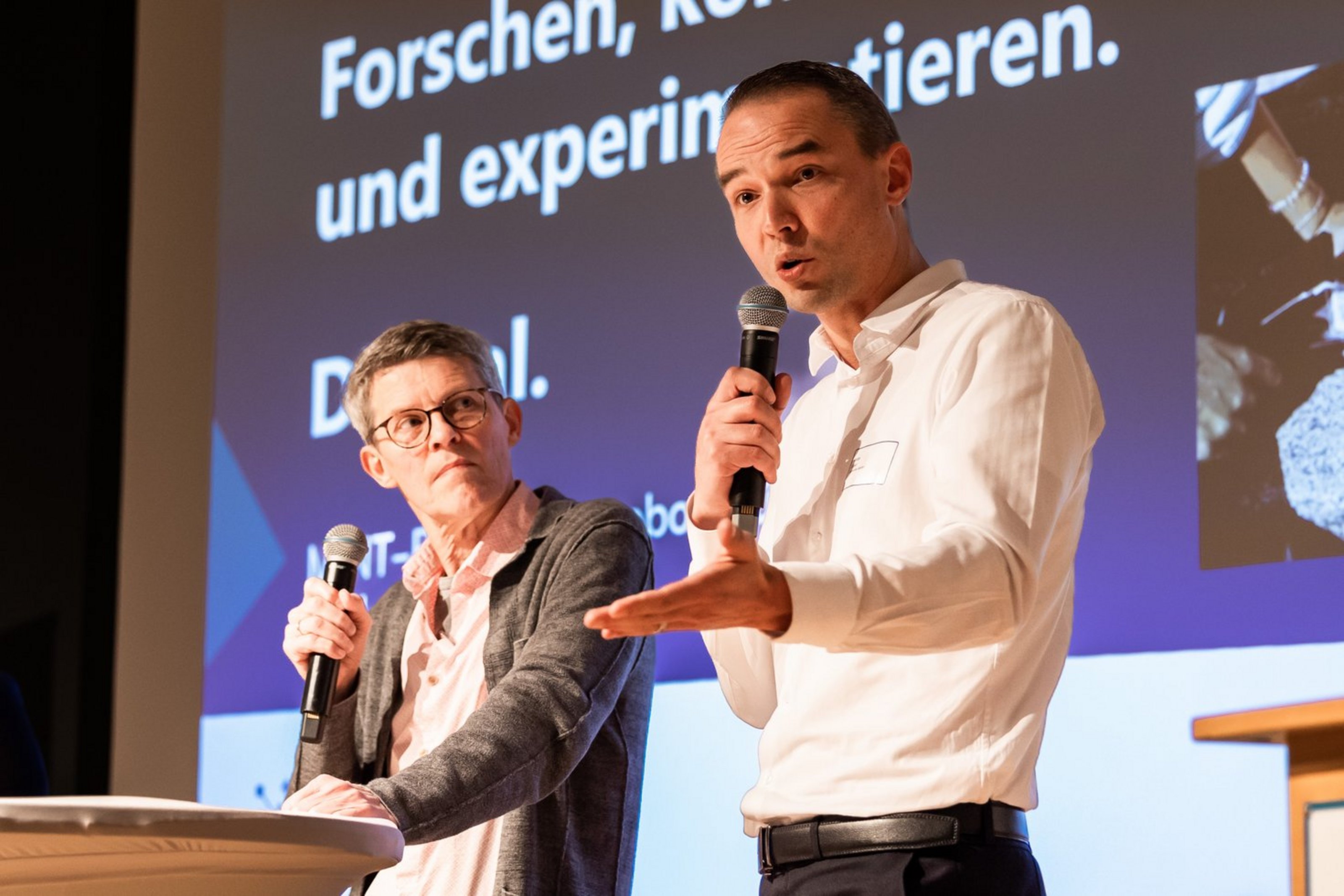 Zwei Männer stehen vor einem Bildschirm, einer spricht in ein Mikrofon und gestikuliert mit der Hand