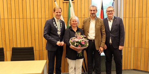 Drei Männer und eine Frau stehen vor einer Wand mit Holfvertäfelung. Hinter ihnen sieht man Flaggen. Die Frau hält einen Blumenstrauß in der Hand.