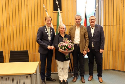 Drei Männer und eine Frau stehen vor einer Wand mit Holfvertäfelung. Hinter ihnen sieht man Flaggen. Die Frau hält einen Blumenstrauß in der Hand.