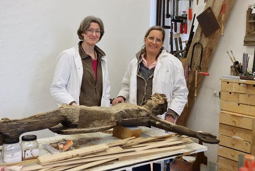 Zwei Restauratorinnen stehen in einer Werkstatt an einem Arbeitstisch. Auf dem Tisch liegt eine stark verwitterte, hölzerne Christusfigur aus einem alten Wegekreuz. Die Frauen tragen weiße Kittel über warmer Kleidung und lächeln in die Kamera. Im Hintergrund sind Werkzeuge wie Sägen, Schraubzwingen und Pinsel ordentlich an der Wand und auf Regalen angebracht. Auf dem Tisch liegen Holzleisten, Restaurierungsmaterialien und Werkzeuge. Die Szene zeigt die handwerkliche Restaurierung historischer Holzskulpturen.