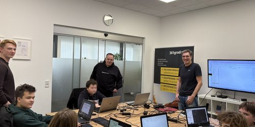 Fünf Jugendliche sitzen an einem Gruppentisch mit Laptops. Daneben stehen drei Männer.