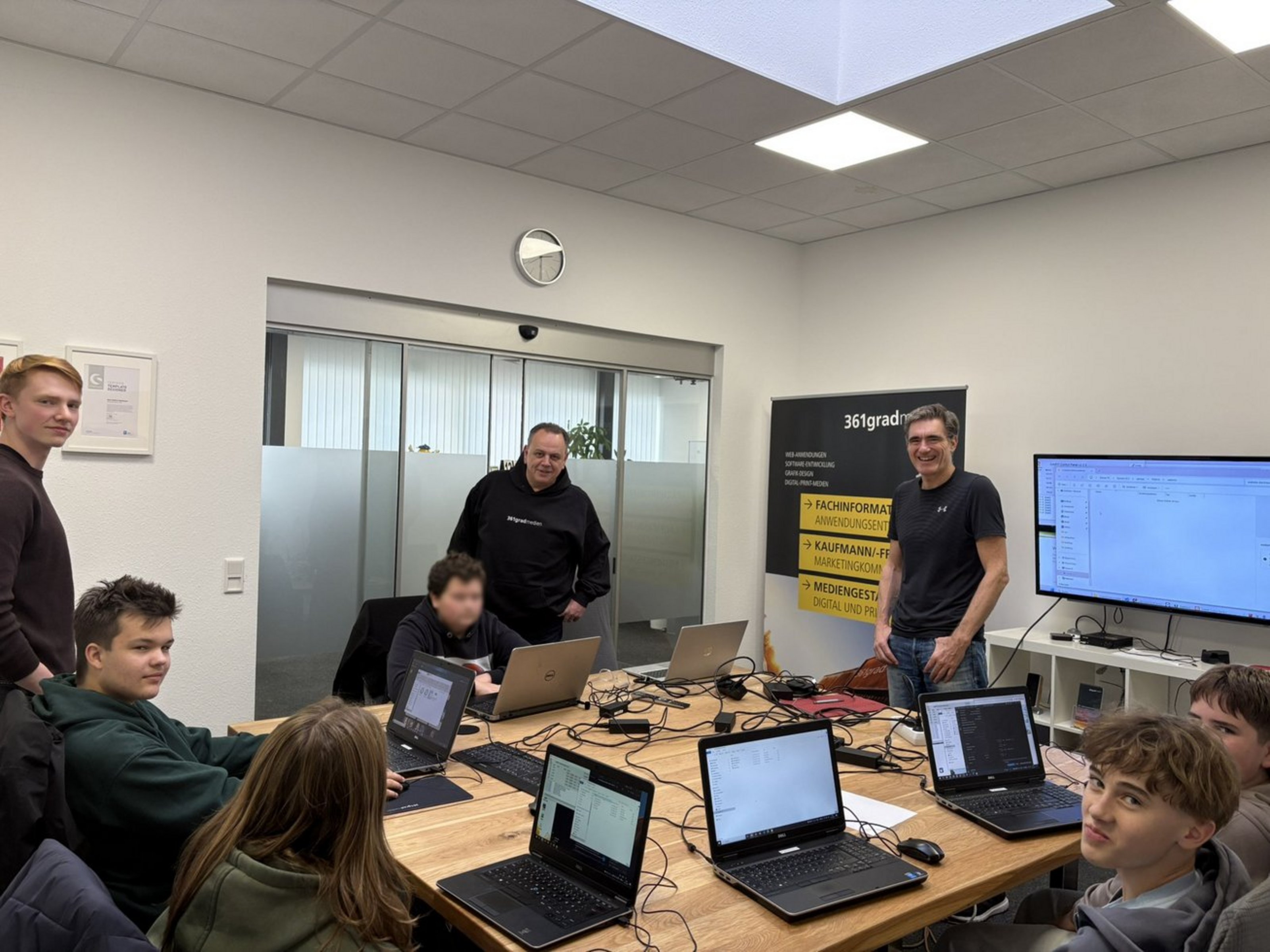 Fünf Jugendliche sitzen an einem Gruppentisch mit Laptops. Daneben stehen drei Männer.