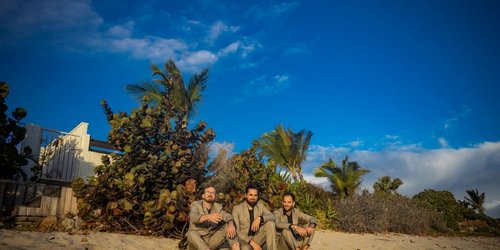 Drei Musiker von Mario Rom’s Interzone sitzen im Sand vor tropischer Vegetation.