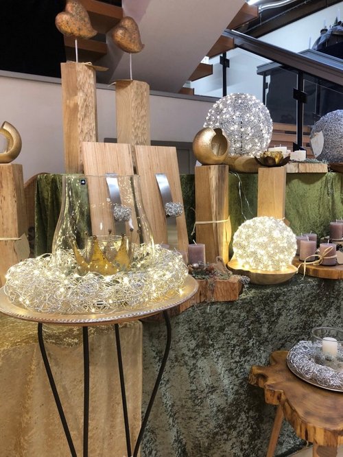 Ein dekorativer Ausstellungstisch mit warmweißen Lichterketten und kunstvollen Objekten: Auf mehreren Holzsockeln stehen goldfarbene Skulpturen und runde, leuchtende Drahtkugeln. Ein großes Glasgefäß mit einer goldenen Krone im Inneren ist von einem Kranz aus feinen, beleuchteten Drähten umgeben.