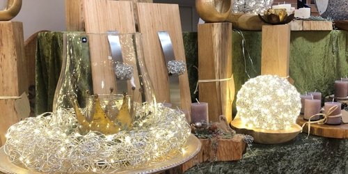Ein dekorativer Ausstellungstisch mit warmweißen Lichterketten und kunstvollen Objekten: Auf mehreren Holzsockeln stehen goldfarbene Skulpturen und runde, leuchtende Drahtkugeln. Ein großes Glasgefäß mit einer goldenen Krone im Inneren ist von einem Kranz aus feinen, beleuchteten Drähten umgeben.