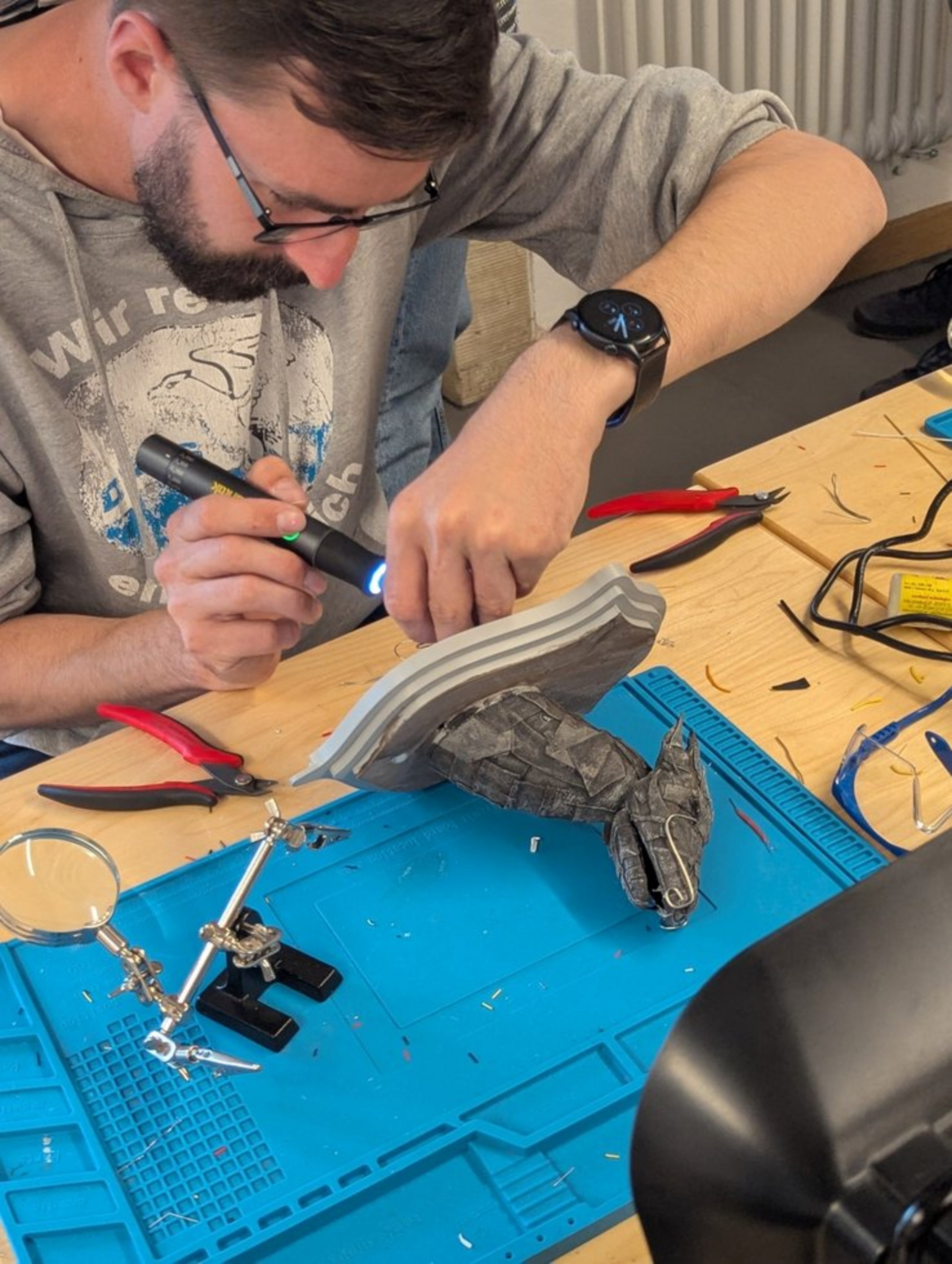 Eine Person mit Bart und Brille hält ein Gerät in der Hand, das er an einem Drachenkopf hält. Ers itzt an einem Tisch mit Unterlage und herum liegenden Kabeln.