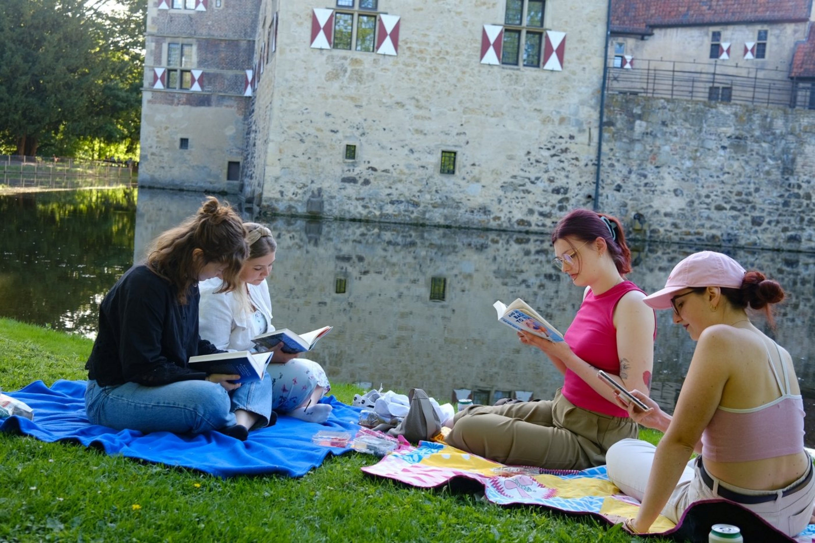 Vier Personen sitzen mit Büchern auf Picknickdecken vor der Burg Vischering und lesen gemeinsam.
