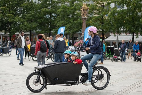 Eine Frau mit blauem Pulli, Jeans und einem rosa Fahrradhelm mit weißen Punkten fährt auf einem Lastenrad. Vorne in der Transportbox des Lastenrades sitzen zwei Kinder mit blauem Fahrradhelm. Sie fährt über einen gepflasterten Platz in einer Stadt. Im Hintergrund sind viele Personen, weitere Lastenräder, die geparkt sind, ein Brunnen, Bäume und Café und Läden zu sehen.