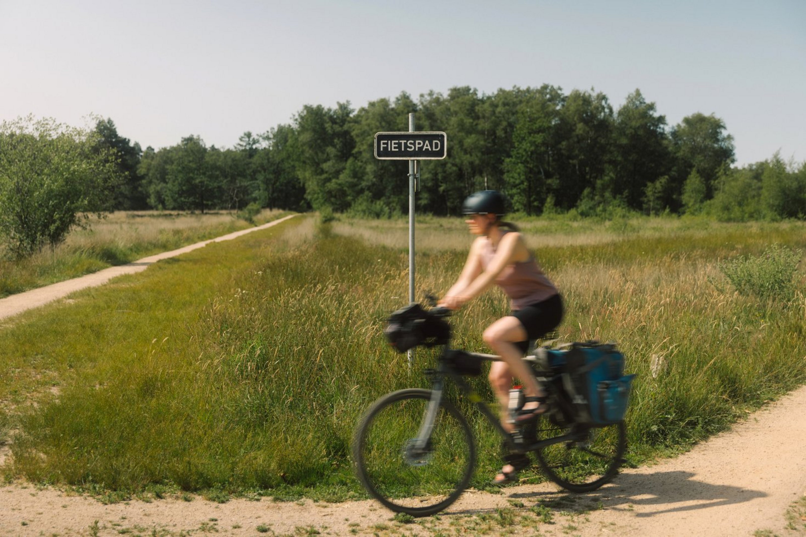 Eine Radfahrerin fährt um die Kurve auf einem Weg. Hinter ihr ist eine Wiese und Wald.
