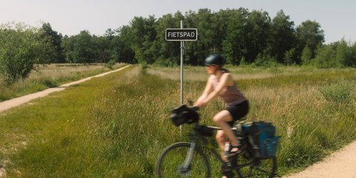 Eine Radfahrerin fährt um die Kurve auf einem Weg. Hinter ihr ist eine Wiese und Wald.