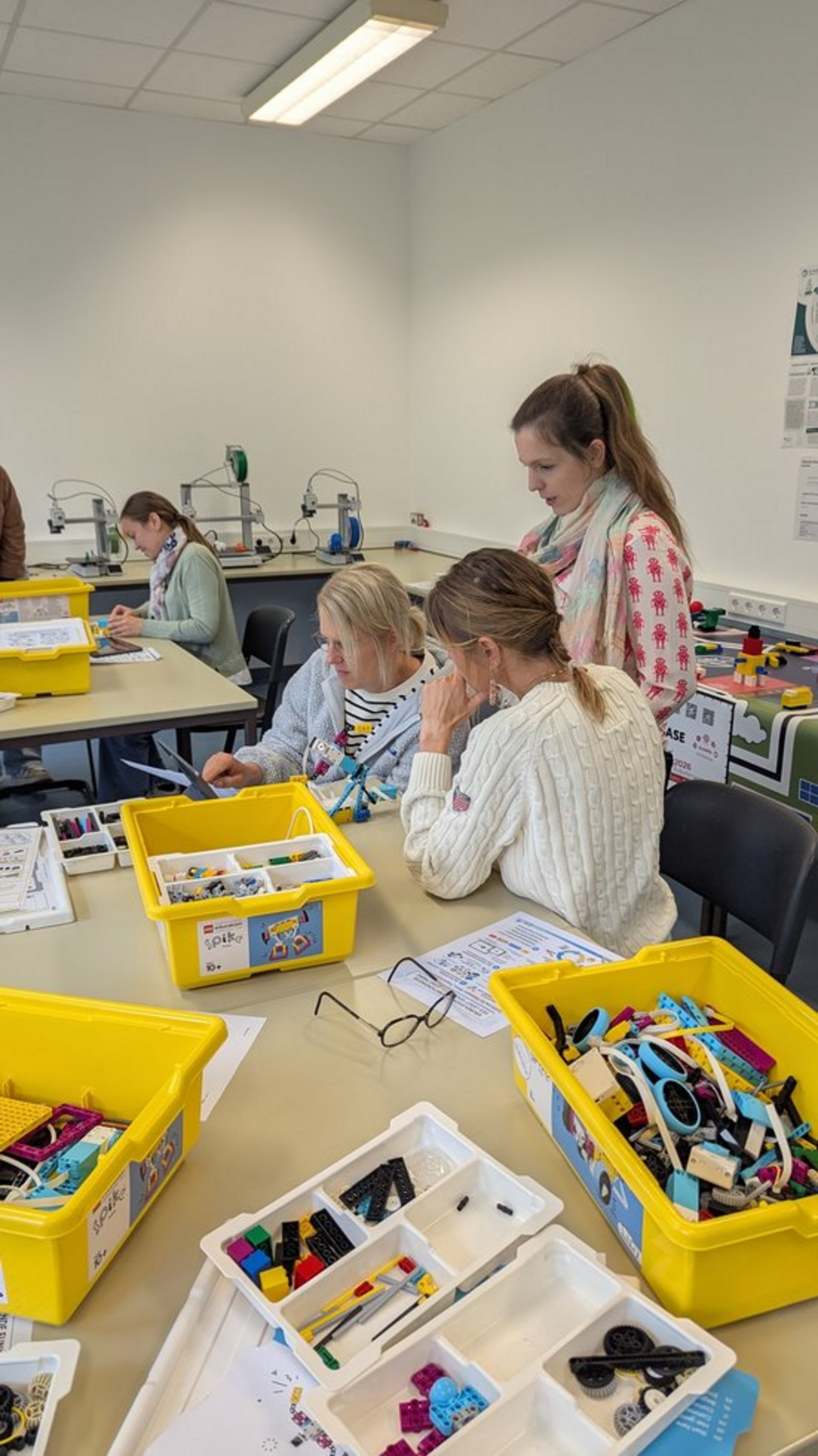 Drei Frauen stehen und sitzen an einem Tisch, auf dem gelbe Kisten mit Bauteilen platziert sind.