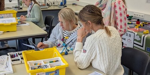 Drei Frauen stehen und sitzen an einem Tisch, auf dem gelbe Kisten mit Bauteilen platziert sind.