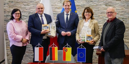 Fünf Personen stehen hinter einem Tisch, auf dem vier Fahnen (von polen, Deutschland, dem Kreis Nysa und Europa). Sie stehen vor einer grauen Wand.