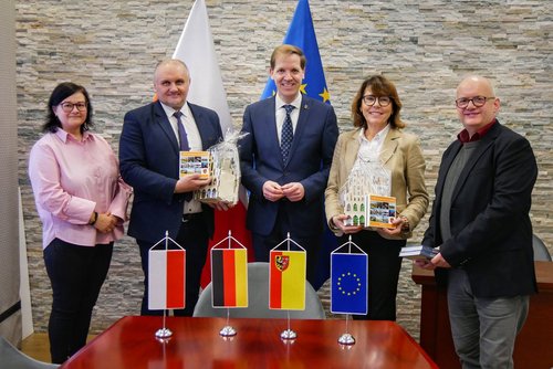 Fünf Personen stehen hinter einem Tisch, auf dem vier Fahnen (von polen, Deutschland, dem Kreis Nysa und Europa). Sie stehen vor einer grauen Wand.