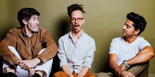 Drei Männer sitzen an eine Wand gelehnt auf dem Boden nebeneinander. Die beiden äußeren Männer schauen zu dem Mann in der Mitte.