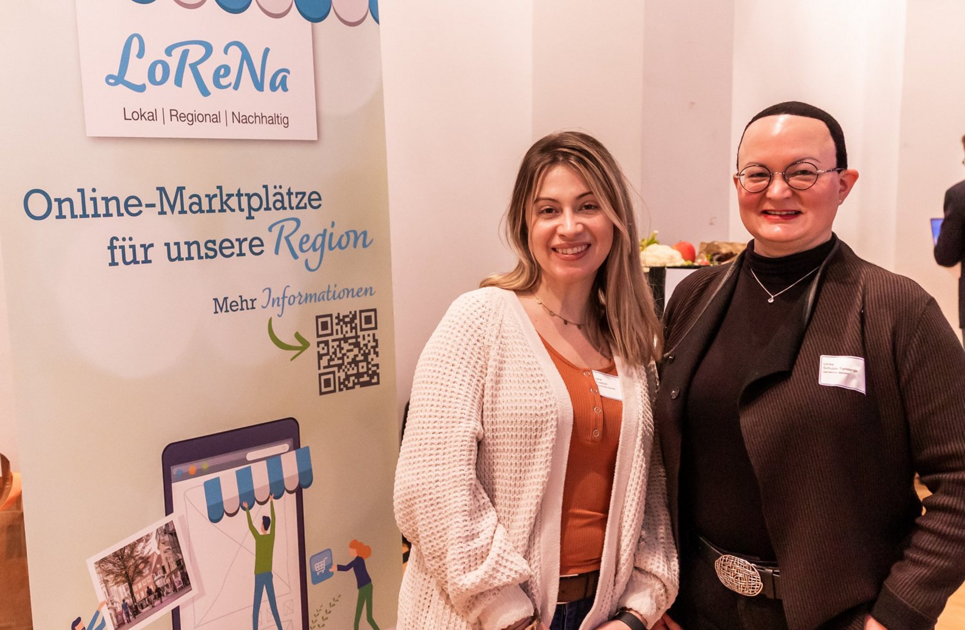 Zwei Frauen stehen nebeneinander vor einem Banner mit dem Schriftzug 'LoReNa Online-Marktplätze für unsere Regionen'