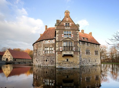 Historische Wasserburg mit runder Hauptburg und rotem Ziegeldach, umgeben von einem Wassergraben. Die Fassade besteht aus Naturstein und Backstein, mit weißen und roten Fensterläden. Vor der Burg spiegelt sich das Gebäude im ruhigen Wasser. Im Hintergrund sind weitere Gebäude mit ähnlicher Architektur und ein bewaldeter Bereich zu sehen. Der Himmel ist teilweise bewölkt mit etwas Sonnenschein.