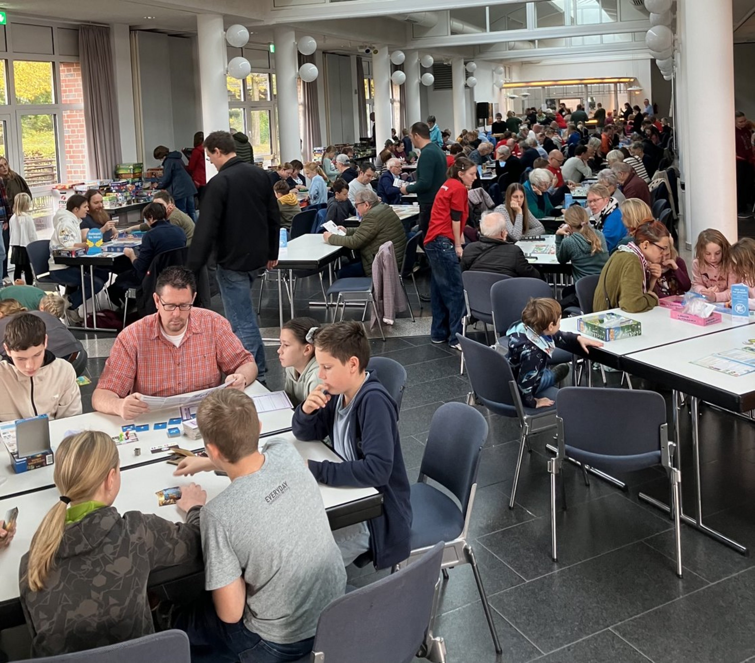 In einem großen offenen Raum sitzen zahlreiche Menschen an Gruppentischen und spielen Spiele.