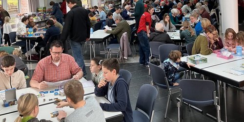 In einem großen offenen Raum sitzen zahlreiche Menschen an Gruppentischen und spielen Spiele.