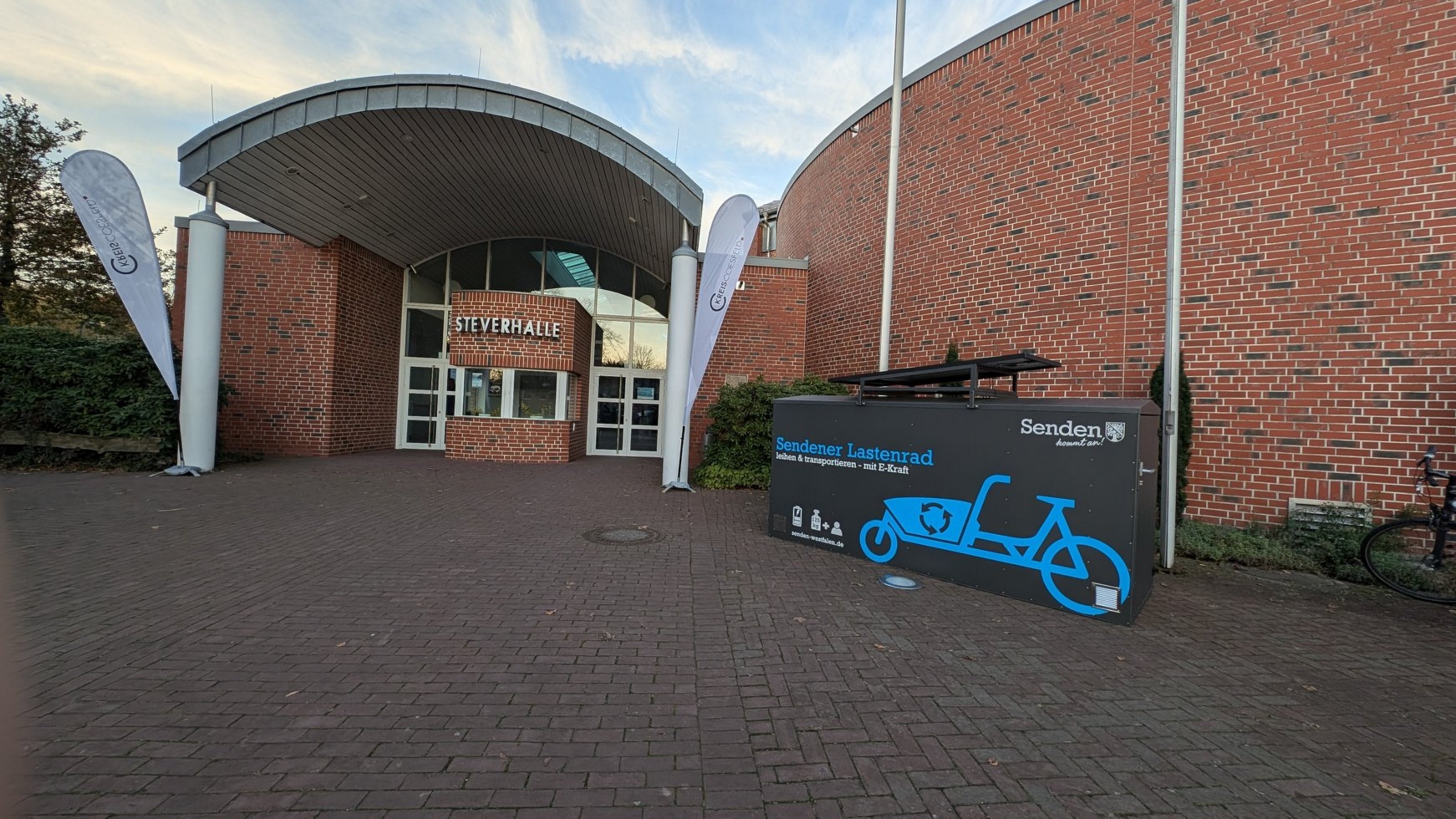 Eingangsbereich der Silverhalle mit zwei weißen Fahnen und einem schwarzen Lastenradständer mit blauer Lastenradgrafik und Schriftzug 'Sendener Lastenrad' vor rotem Backsteingebäude.