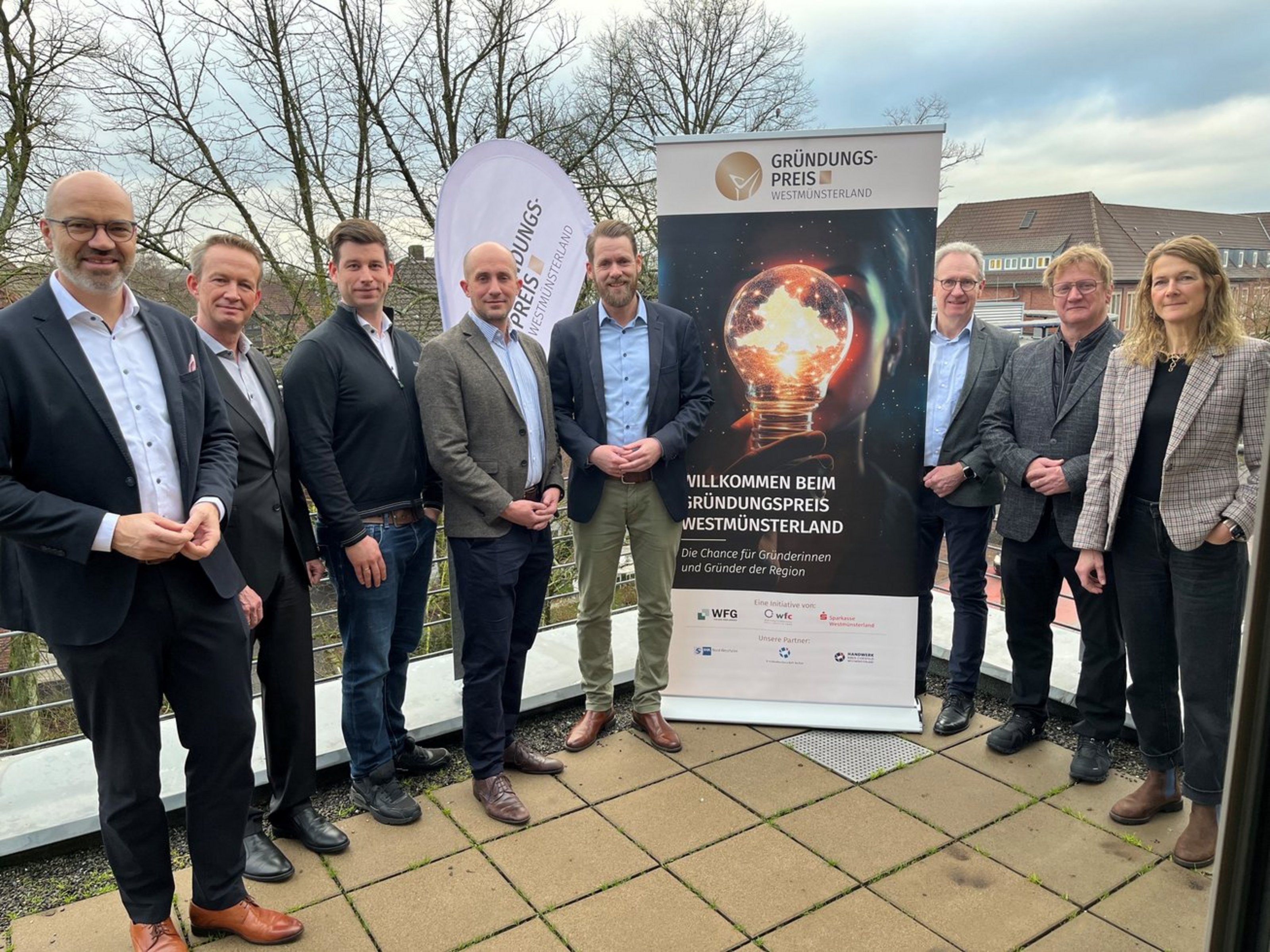Acht Personen, darunter eine Frau und sieben Männer, stehen nebeneinander auf einem Balkon. Zwischen ihnen steht ein BAnner mmit der Aufschrift "Gründungspreis Westmünsterland" und zeigt eine Glühbirne.