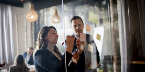Eine Frau und ein Männ stehen vor einer Glasscheibe, an die Post-its geklebt sind. Beide tragen ein Jacket. Die Frau hält einen Stift in der Hand. Im Hintergrund sitzen Personen an einem Tisch.