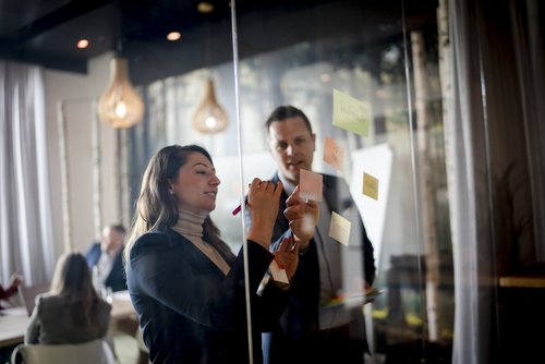 Eine Frau und ein Männ stehen vor einer Glasscheibe, an die Post-its geklebt sind. Beide tragen ein Jacket. Die Frau hält einen Stift in der Hand. Im Hintergrund sitzen Personen an einem Tisch.