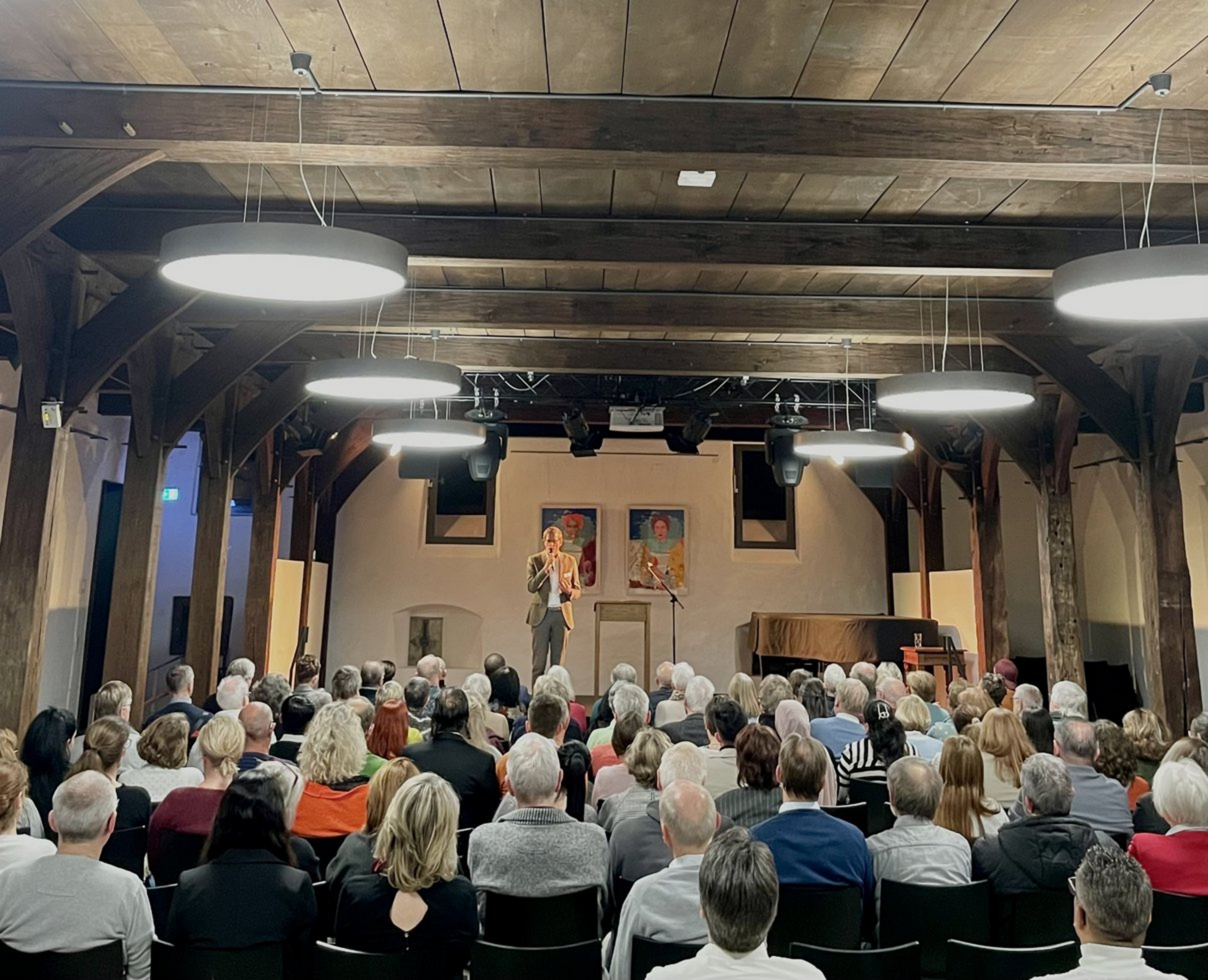 In einem Saal sitzen viele Menschen im Publikum und schauen Richtung Bühne, auf der eine Person im Anzug steht.