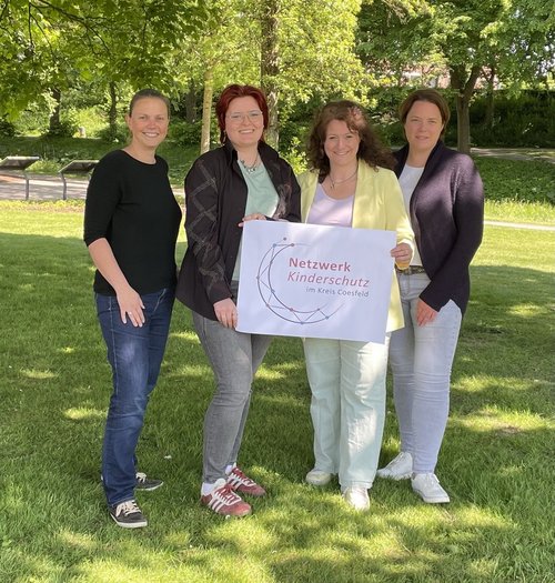 Vier Frauen stehen auf einer Rasenfläche und präsentieren ein Schild, auf dem das neue Logo des Netzwerks Kinderschutz zu sehen ist. Das Logo setzt sich aus einem doppeltel dreiviertelkreis zusammen, der mit bunten Punkten versehen ist, die mit Linien verbunden sind. Im Logo steht "Netzwerk Kinderschutz im Kreis Coesfeld".