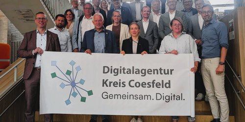 Eine Gruppe von Männern und Frauen steht auf einer Treppe. Sie halten ein großes Schild mit der Aufschrift Digitalagentur Kreis Coesfeld.