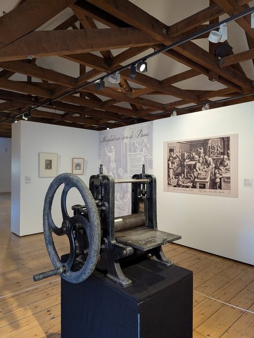 Ein Raum in einem Museum mit Holzboden und Holzbalkendecke. Im Vordergrund steht eine historische Druckpresse mit großem Handrad auf einem schwarzen Sockel. Im Hintergrund sind weiße Ausstellungswände mit gerahmten Drucken und einer großen Abbildung einer Werkstattszene zu sehen. Eine Wand trägt den Schriftzug „Magdalena van de Passe“.