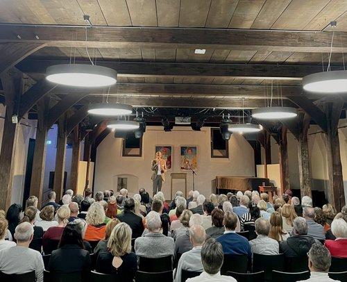 In einem Saal sitzen viele Menschen im Publikum und schauen Richtung Bühne, auf der eine Person im Anzug steht.