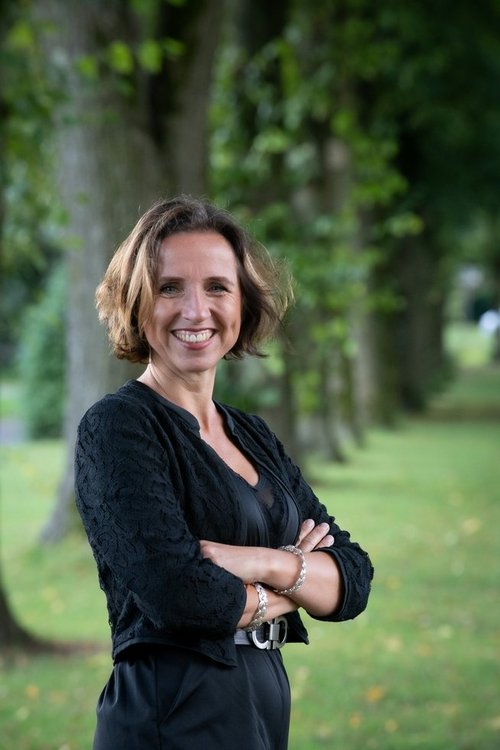 Porträt einer Frau mit schulterlangem, braunem Haar, die freundlich in die Kamera lächelt. Sie trägt eine schwarze Bluse mit Spitzenmuster und hat die Arme vor der Brust verschränkt. Im Hintergrund ist eine grüne Parklandschaft mit Bäumen zu sehen, die leicht unscharf ist. 