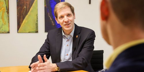 Landrat Dr. Christian Schulze Pellengahr sitzt an einem Tisch und schaut eine weitere Person an, die die Kamera mit dem Rücken auffängt.