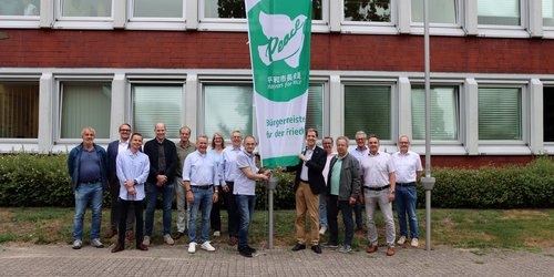 Eine Gruppe von Männern und Frauen stehen vor einem Gebäude um einen Fahnenmast herum. Die Fahne ist grün und weiß. Auf ihr sieht man eine Taube mit dem Wort Peace sowie die Beschriftung Mayors for Peace und Bürgermeister für den Frieden.