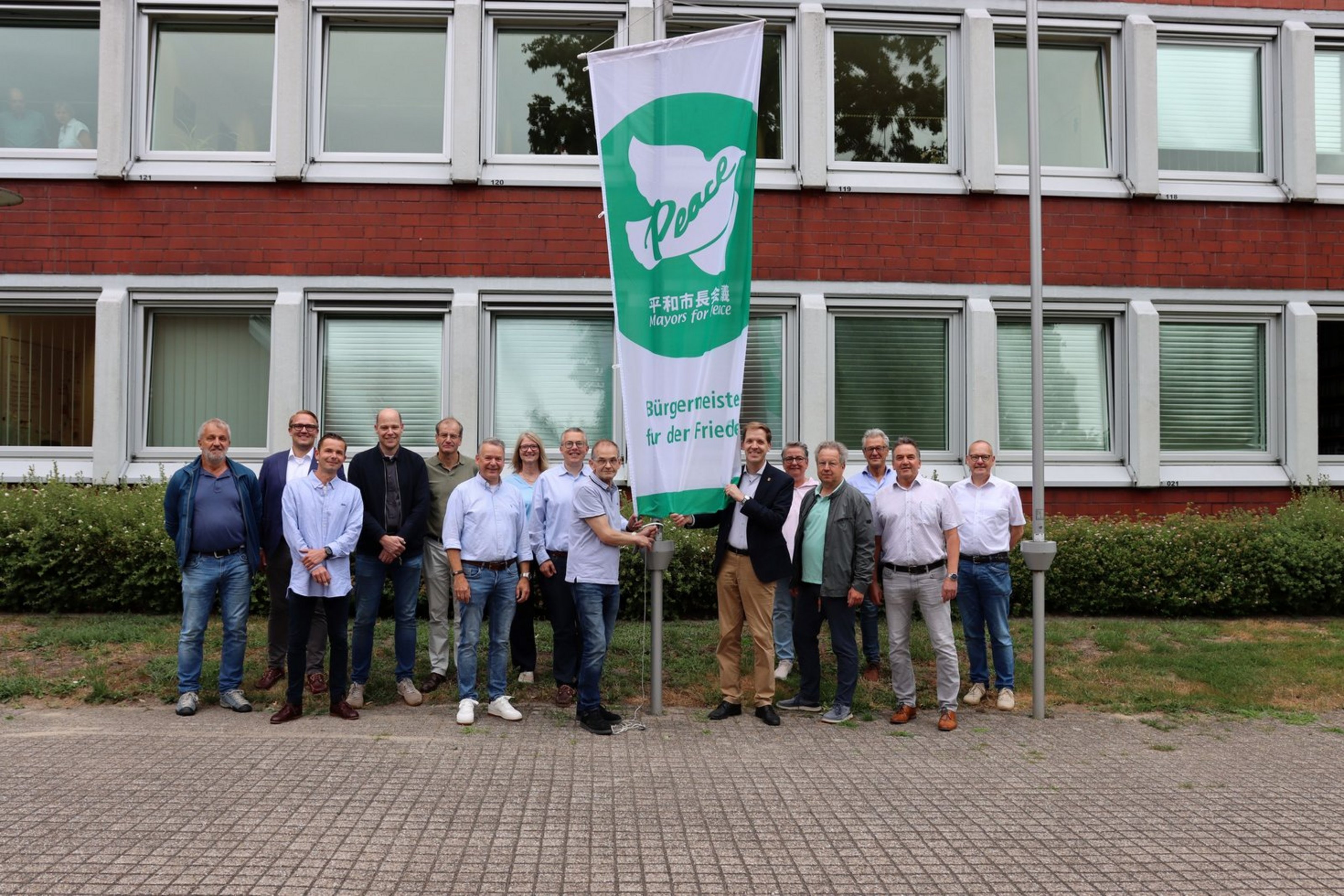 Eine Gruppe von Männern und Frauen stehen vor einem Gebäude um einen Fahnenmast herum. Die Fahne ist grün und weiß. Auf ihr sieht man eine Taube mit dem Wort Peace sowie die Beschriftung Mayors for Peace und Bürgermeister für den Frieden.