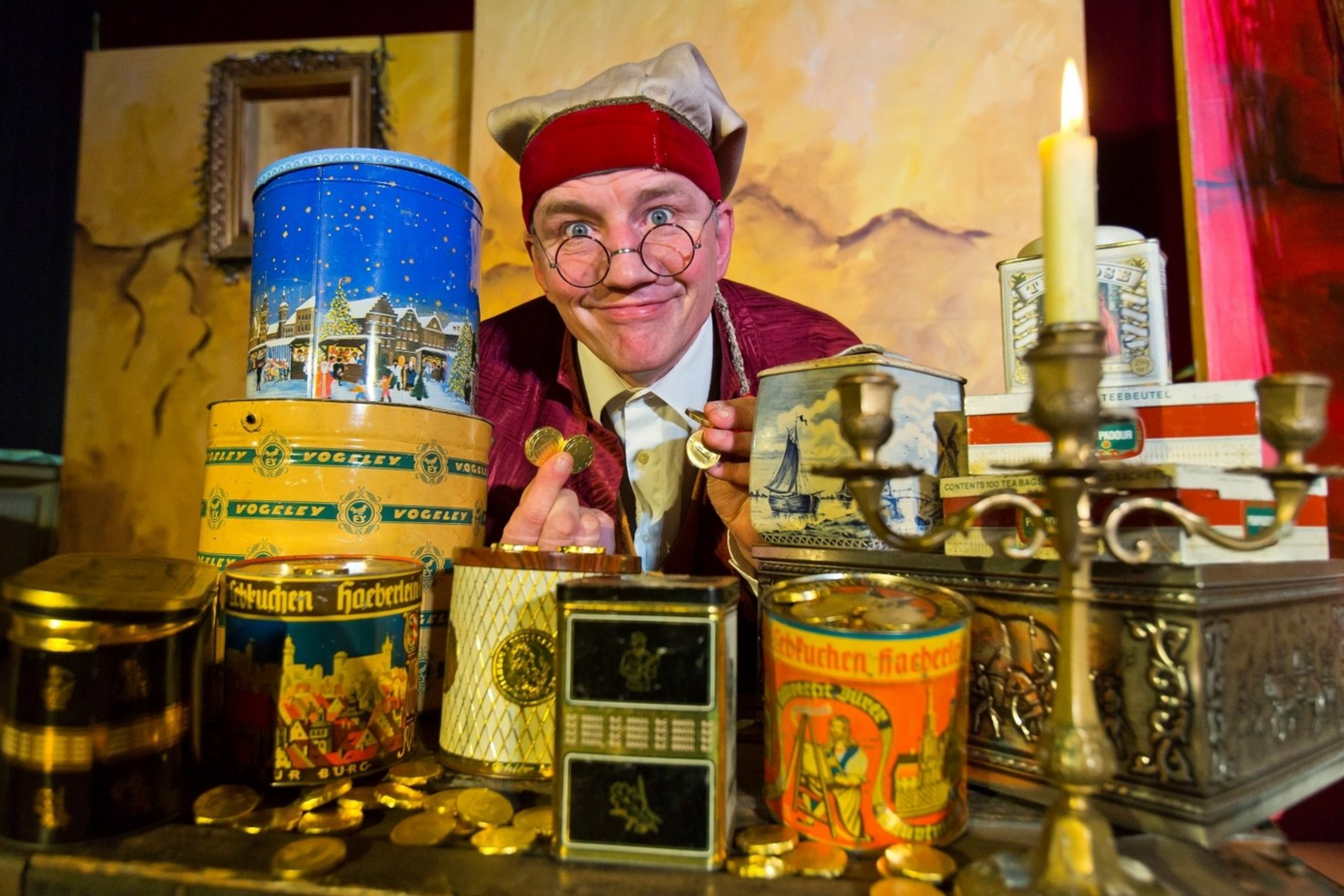 Ein Schauspieler in altertümlicher Kleidung und Mütze sitzt vor vielen bunten Blechdosen und hält lachend goldene Münzen in den Händen.