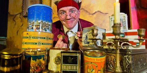 Ein Schauspieler in altertümlicher Kleidung und Mütze sitzt vor vielen bunten Blechdosen und hält lachend goldene Münzen in den Händen.