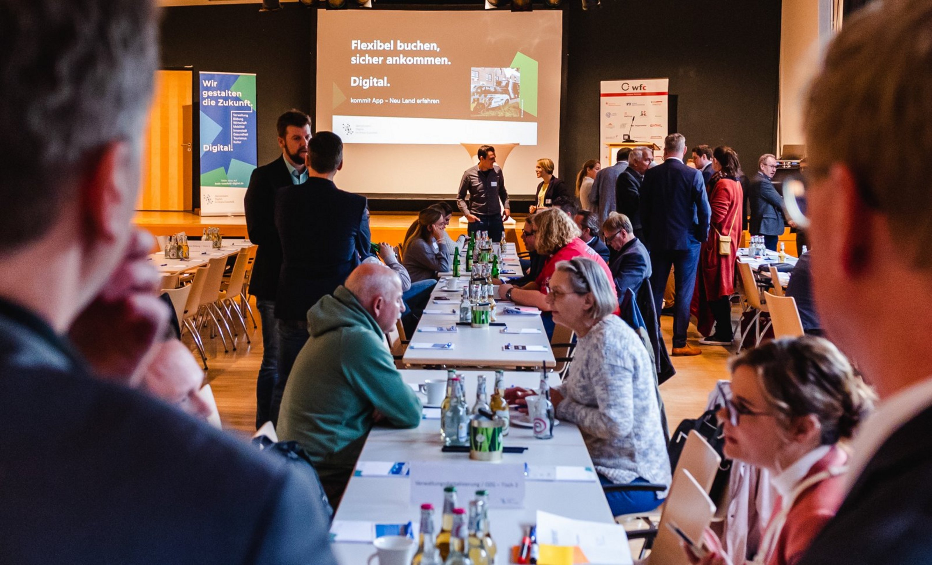 Teilnehmende einer Fachveranstaltung sitzen und stehen an mehreren Tischen im Austausch. Im Hintergrund ist eine Präsentation zu einem digitalen Zukunftsthema zu sehen.