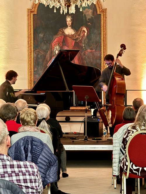 Vor Publikuzm spielen zwei Männer auf einer Bühne Piano und Bass. Hinter ihnen an der Wand hängt ein großes Gemälde von einer historischen Person.