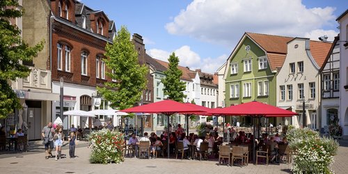 Eine Marktplatz umgeben von älteren Häusern, auf dem in der Mitte mehrere Sonnenschirme und Sitzgelegenheiten sich befinden.