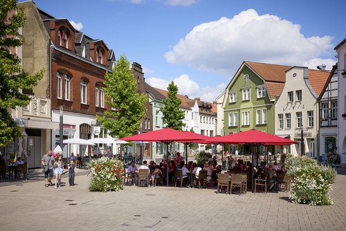 Eine Marktplatz umgeben von älteren Häusern, auf dem in der Mitte mehrere Sonnenschirme und Sitzgelegenheiten sich befinden.