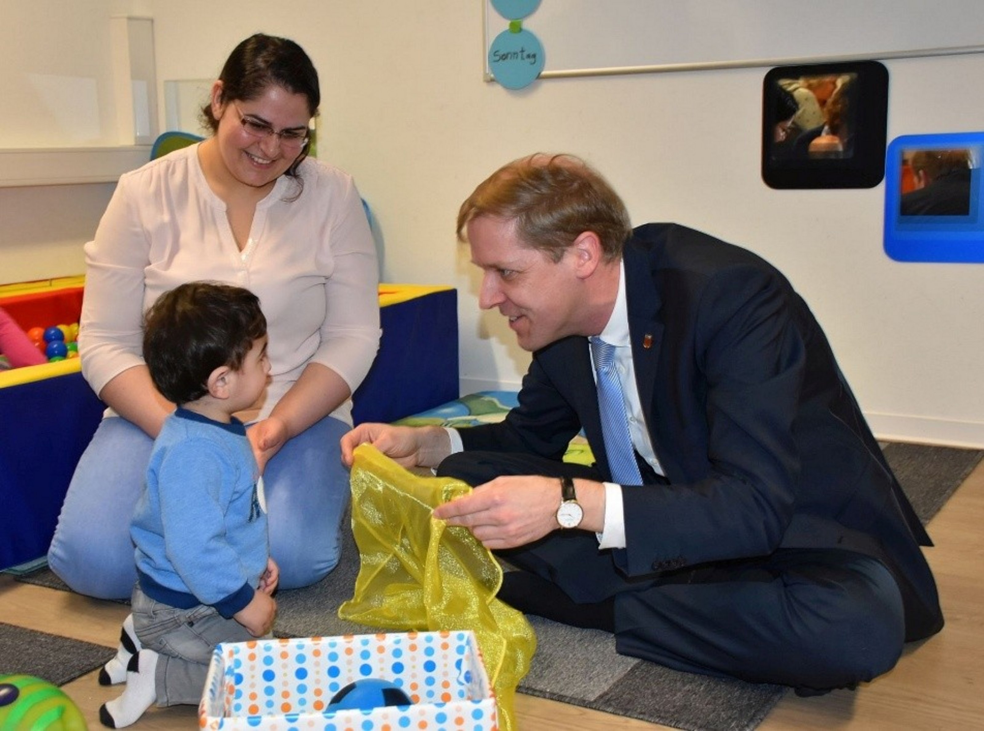 Saeruz Sandy Culu sitzt mit ihrem Sohn in einem Spielraum, während Landrat Dr. Christian Schulze Pellengahr sich auf Augenhöhe dem Kind zuwendet und mit einem Spieltuch interagiert.