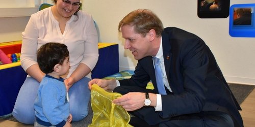 Saeruz Sandy Culu sitzt mit ihrem Sohn in einem Spielraum, während Landrat Dr. Christian Schulze Pellengahr sich auf Augenhöhe dem Kind zuwendet und mit einem Spieltuch interagiert.