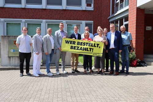Mehrere Personen stehen vor dem Kreishaus und halten ein großes gelbes Banner mit der Aufschrift „Wer bestellt, bezahlt!“ in die Kamera.