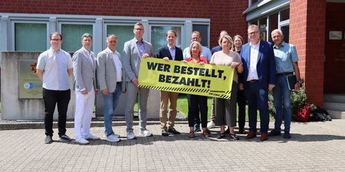 Mehrere Personen stehen vor dem Kreishaus und halten ein großes gelbes Banner mit der Aufschrift „Wer bestellt, bezahlt!“ in die Kamera.