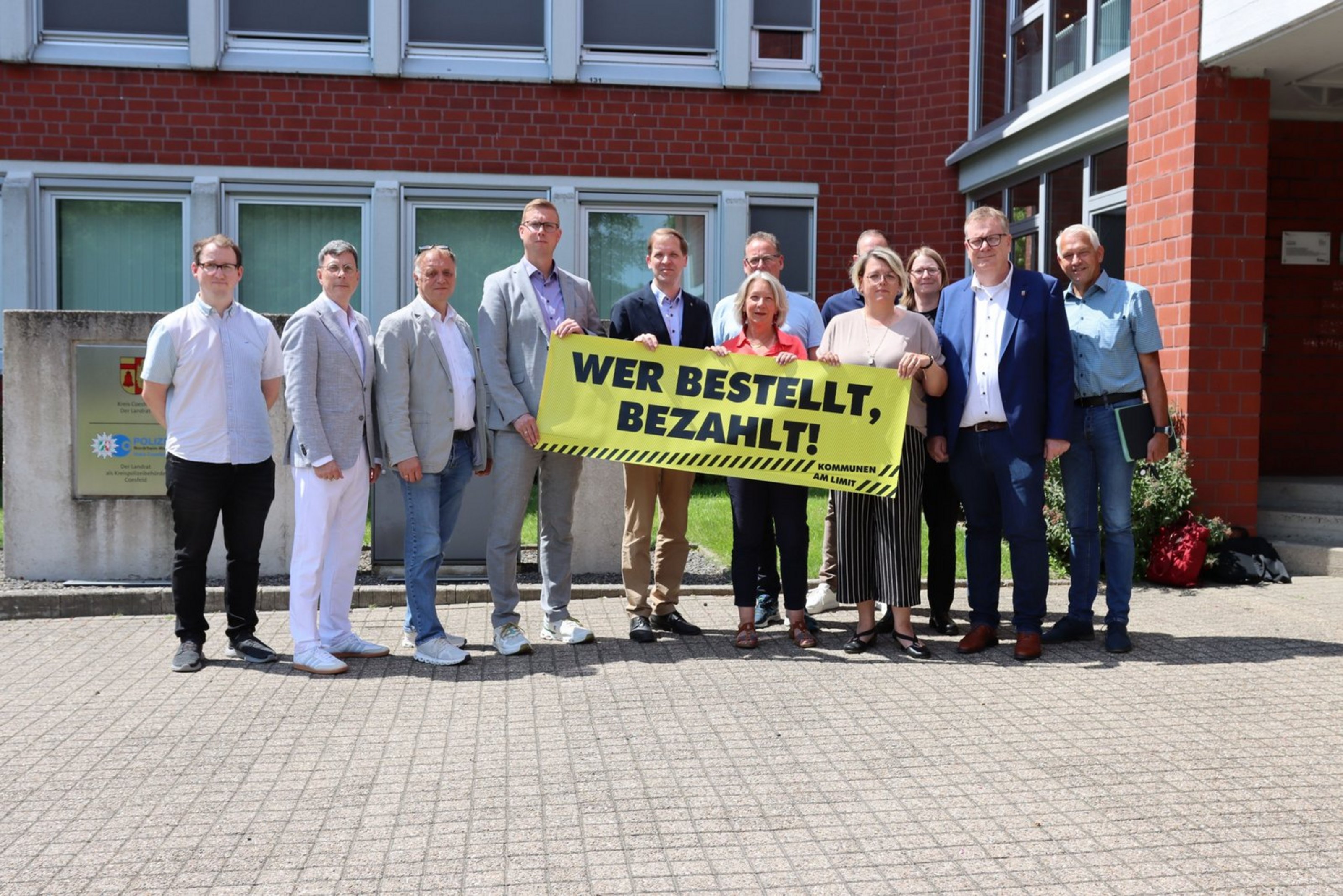 Mehrere Personen stehen vor dem Kreishaus und halten ein großes gelbes Banner mit der Aufschrift „Wer bestellt, bezahlt!“ in die Kamera.